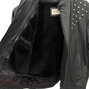 以下が含まれることがあります： 黒い革のジャケット。ジッパー開閉で、内側は黒い裏地。肩には銀色のスタッズ、襟には「CHO」の文字が入った小さなラベルが付いています。