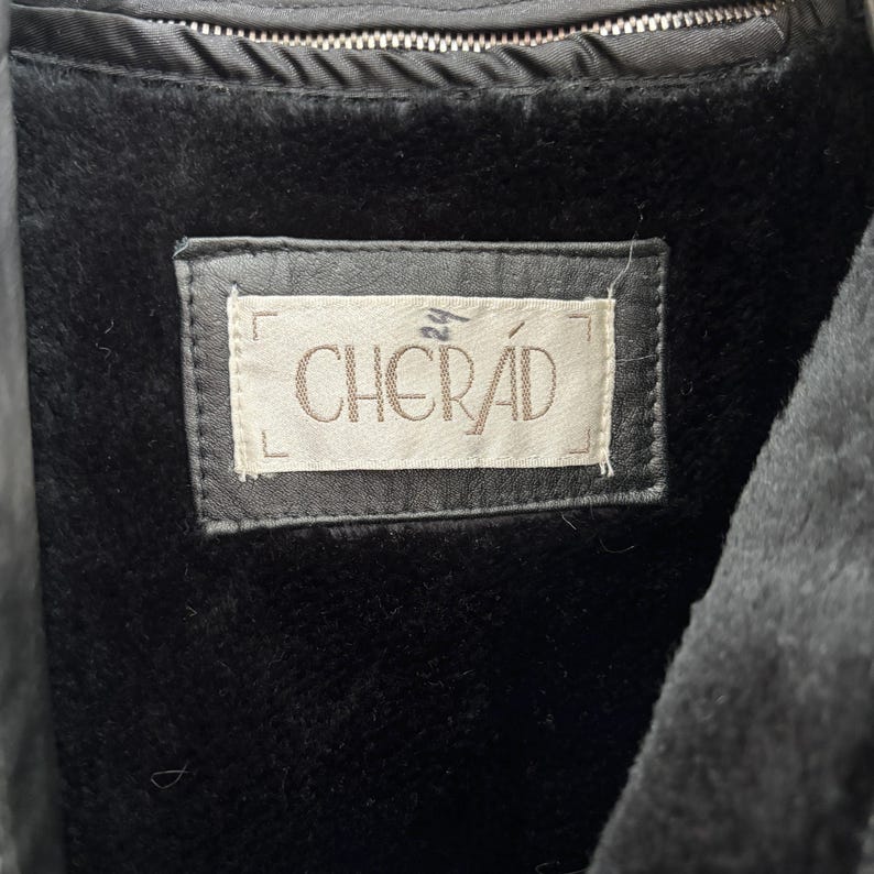 以下が含まれることがあります： 長方形のフレームに「CHERAD」と書かれたラベルが付いた黒いコートの内側のクローズアップ。コートは黒色の豪華な裏地と上部にジッパーが付いています。ラベルには数字の「24」も印刷されています。