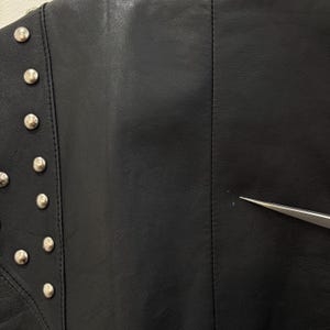 以下が含まれることがあります： 左側に銀色の金属スタッドが付いた黒い革のジャケットのクローズアップ。 ジャケットの中央には垂直の縫い目があり、右側には小さなマークがあります。