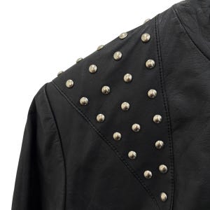 以下が含まれることがあります： 肩に銀色の金属スタッドが付いた黒い革のジャケットのクローズアップ。 ジャケットはテーラードフィットのクラシックなデザインで、スタッドは均等に配置されています。