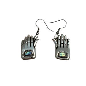 May include: A pair of silver-toned earrings shaped like open hands. Each earring features a small, oval-shaped iridescent stone set in the palm. The earrings have a hook closure for pierced ears, and are displayed against a white background.