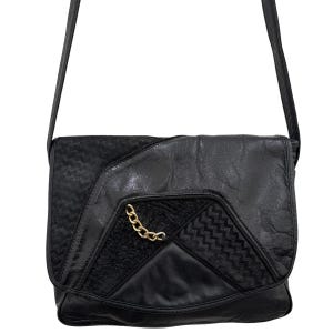 May include: A black shoulder bag featuring a flap with a mix of smooth and textured panels. The design includes sections with a chevron-like pattern and a small gold-toned chain link accent attached to the front flap. It has a long shoulder strap.