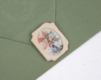 May include: An olive green envelope with a floral wax seal. The seal is an oval shape with a cream-coloured border and a watercolour floral design. The envelope has a monogram in the upper left corner.