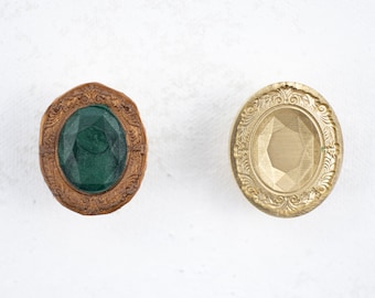 May include: Two oval-shaped decorative knobs with ornate gold frames. One knob features a green gemstone, while the other has a clear glass gemstone.