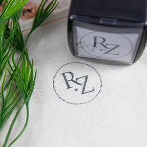 May include: A black self-inking stamp with the initials "RZ" in a circle. The stamp is on a white fabric pouch with the same initials stamped in the center.