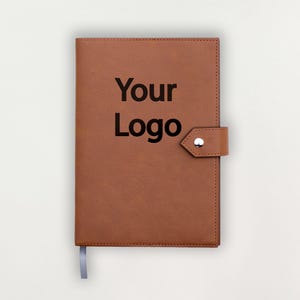 Peut inclure: Un journal en cuir marron avec le texte "Your Logo" en noir. Le journal a une fermeture à lanière avec un bouton argenté et un signet en ruban gris. La couverture est marron unie et texturée.