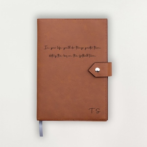 May include: A brown leather journal with a snap closure and a grey ribbon bookmark. The journal features the text "In your life you'll do things greater than dating the boy on the football team" and initials "TS" on the cover.