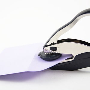 May include: A silver and black self-inking stamp with a handle. The stamp is being used to stamp a light purple envelope.