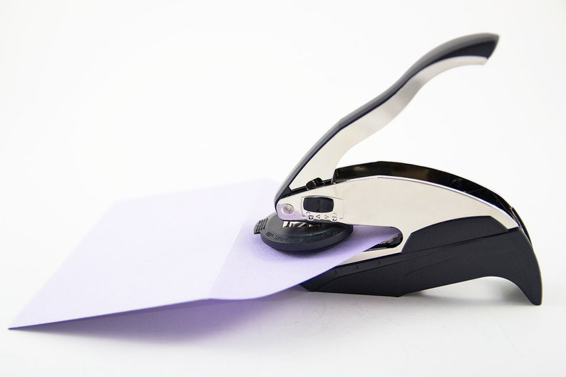May include: A silver and black self-inking stamp with a handle. The stamp is being used to stamp a light purple envelope.