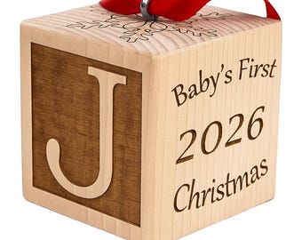 Könnte beinhalten: Ein helles Holzblock-Ornament mit einem großen eingravierten Buchstaben J auf der Vorderseite. Die Seite ist mit dem Text Baby's First 2026 Christmas graviert. Auf der Oberseite befindet sich eine Schneeflocke und ein rotes Band mit Metallring.