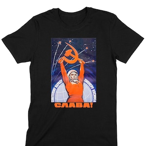 Peut inclure: T-shirt noir avec un poster de propagande spatiale soviétique vintage représentant un cosmonaute tenant un marteau et une faucille devant un ciel étoilé. Le poster est écrit en russe et dit "Pionnier de l'espace" et "Gloire au peuple soviétique !".