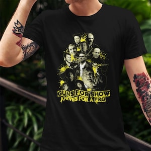 Peut inclure: T-shirt noir avec un motif jaune représentant plusieurs visages et le texte "GUNS FOR SHOW KNIVES FOR A PRO". Le motif comprend des éclaboussures de peinture jaune. Le t-shirt a un col rond.