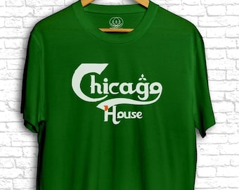 Puede incluir: Una camiseta verde con el texto blanco "Chicago House" en una fuente estilizada. La camiseta está colgada en una percha de madera contra una pared de ladrillos blancos. El texto está en cursiva.