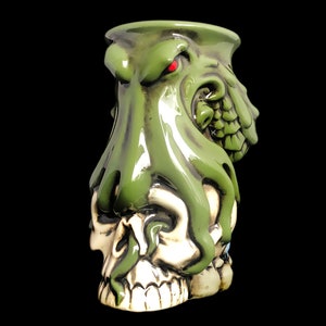 May include: Green ceramic mug shaped like an octopus with a skull face. The octopus has a red eye and is wrapped around the skull.