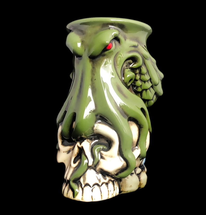 May include: Green ceramic mug shaped like an octopus with a skull face. The octopus has a red eye and is wrapped around the skull.