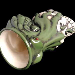 May include: Green and white ceramic mug with a detailed design of a creature with a skull-like face. The mug is tilted to show the inside of the mug.
