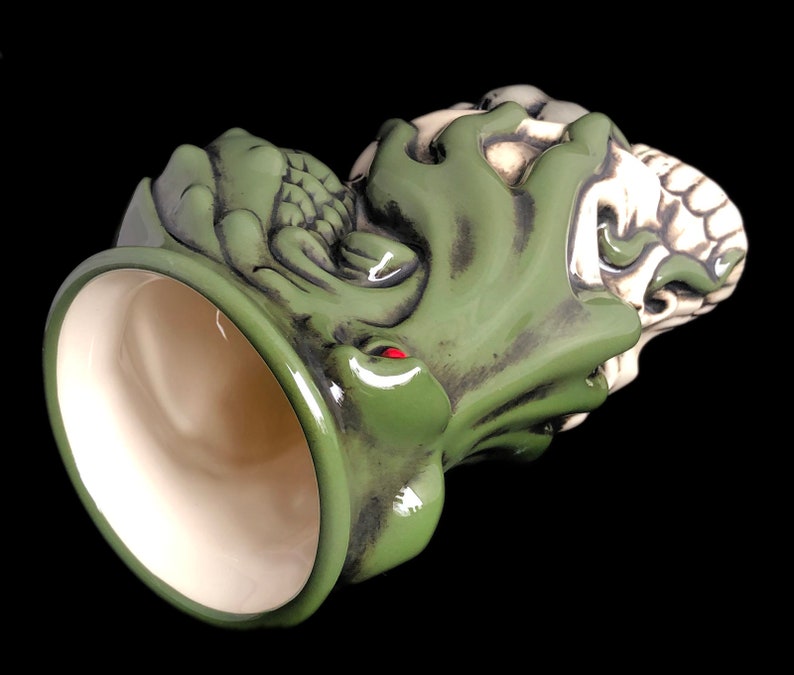 May include: Green and white ceramic mug with a detailed design of a creature with a skull-like face. The mug is tilted to show the inside of the mug.