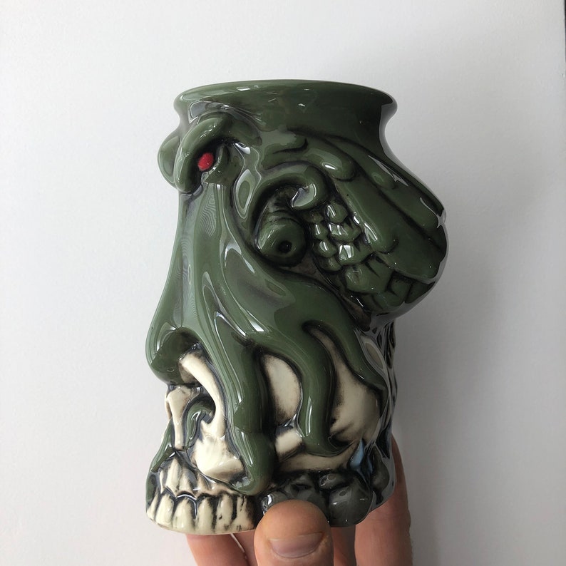 May include: A green ceramic mug shaped like a skull with a green octopus tentacle draped over it. The octopus has a red eye.