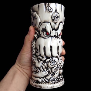 May include: White ceramic tiki mug with a detailed design of a skull-faced octopus with red eyes. The mug has a textured surface and is holding a skull in its tentacles.