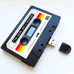 Könnte beinhalten: Ein schwarzer USB-Stick in Form einer Kassette mit dem Text "I've made you a Mixtape" auf der Vorderseite. Der Stick hat ein rot-orange-gelbes Streifenmuster und den Text "90" oben rechts.