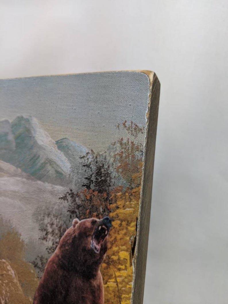 May include: A close-up of a wooden wall art piece featuring a brown bear with its mouth open, roaring. The bear is standing in a forest setting with mountains in the background. The art piece has a rustic, distressed look.