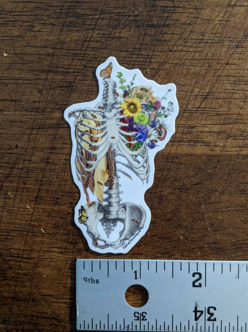 May include: A white sticker with a detailed illustration of a human skeleton with a bouquet of colorful flowers and bees inside the rib cage. The sticker is approximately 2 inches tall and has the text "Tatte DecoArt" at the bottom.