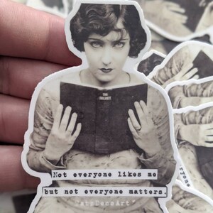 Pu&ograve; includere: Sticker fotografico in bianco e nero di una donna che tiene un libro. Il testo sullo sticker recita "Not everyone likes me but not everyone matters. TatsDecoArt."