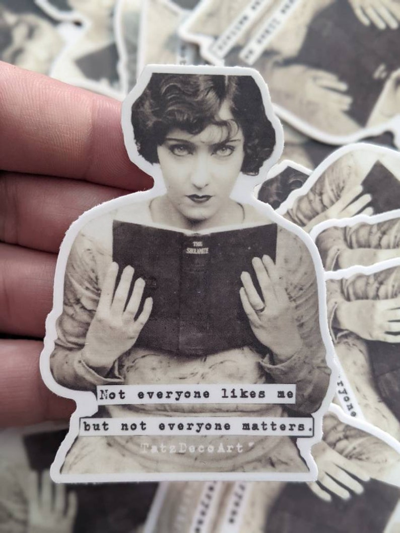 Pu&ograve; includere: Sticker fotografico in bianco e nero di una donna che tiene un libro. Il testo sullo sticker recita "Not everyone likes me but not everyone matters. TatsDecoArt."