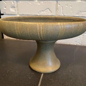 May include: A pedestal bowl with a speckled olive green finish and a wide, shallow basin. The exterior of the bowl features vertical ribbed detailing around the circumference, supported by a sturdy, flared circular base stand.