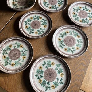 May include: A set of six ceramic plates and one bowl featuring a central textured brown circle surrounded by a wreath of green leaves and yellow flowers. Each piece has a brown rim with a repeating vertical line pattern along the edge.