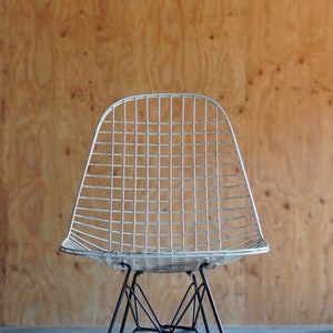 May include: A white wire chair with a black metal base. The chair is in front of a wood panel background.