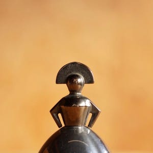 May include: A metallic, dark-toned figurine featuring a stylized human form with hands placed on the hips. The figure wears a prominent, semi-circular headpiece and a wide, bell-shaped skirt, showcasing a smooth, reflective surface throughout.
