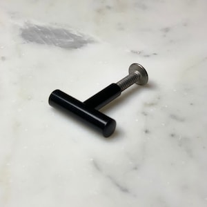 May include: A black cabinet knob with a cylindrical handle and a silver screw. The knob is lying on a white and gray marble surface. The handle is approximately 3 inches long.
