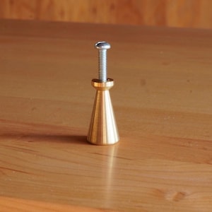 May include: A gold-toned metal cabinet knob with a tapered design and a silver screw.