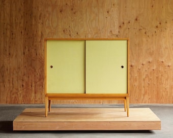 May include: A mid-century modern cabinet with sliding doors. The cabinet has a light wood frame and legs, with pale yellow sliding doors. The cabinet is set on a wooden platform against a wood-paneled wall. The cabinet is approximately 30 inches wide.