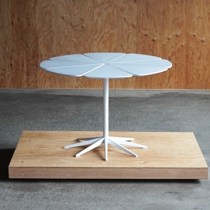 May include: White round table with a unique petal-shaped top and a white, multi-pronged base. The table is on a wooden platform.
