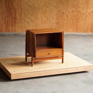 May include: A mid-century modern style wooden nightstand with a single drawer and open shelf. The nightstand is a warm brown color and sits on a light-colored wooden platform. The background is a light brown wooden wall.