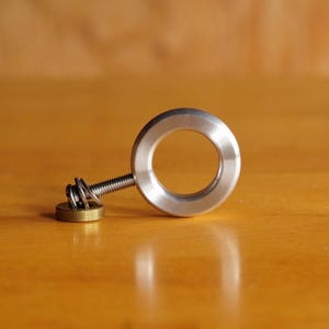 May include: A silver-colored metal ring with a smooth, rounded profile attached to a threaded metal bolt. The bolt is connected to a small, flat, circular brass-toned base piece, creating a mechanical assembly with a distinct industrial appearance.