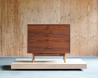 May include: A four-drawer dresser made of dark wood with a simple design. The dresser has a slightly angled base and sits on a wooden platform.