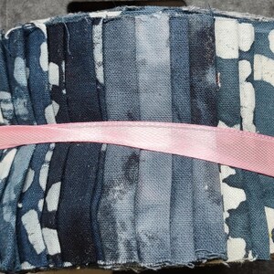 May include: A fabric jelly roll bundle featuring a variety of blue, navy, and grey cotton strips with assorted patterns including dots, mottled textures, and solid shades, all secured together by a single horizontal light pink ribbon.