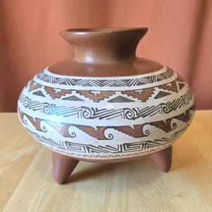 Pode incluir: Um vaso de cerâmica castanho com corpo arredondado e gargalo alargado, assente em três pernas curtas. O vaso é decorado com padrões geométricos em castanho e preto sobre um fundo branco. O vaso tem cerca de 15 cm de altura.