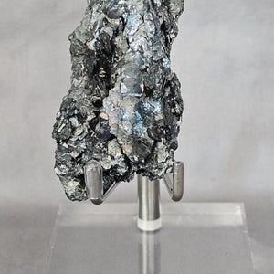 May include: A dark, metallic mineral specimen with a rough, crystalline texture and iridescent blue and silver highlights. The specimen is mounted on a clear acrylic base with a silver-toned metal support stand featuring two prongs.
