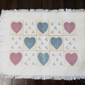 May include: Cream-colored rectangular placemat with a grid pattern of hearts and floral accents. The hearts alternate between dusty rose and blue-gray. The edges are finished with a fringe.
