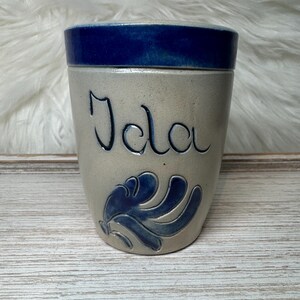 May include: A ceramic mug with a light gray base and a dark blue rim. The name "Ida" is written in cursive on the front, with a blue floral design below. The mug sits on a light wood-grain surface.