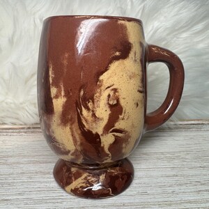 May include: A ceramic mug featuring a marbled pattern of dark brown and cream colors. The mug has a smooth, glossy finish, a sturdy handle on the right side, and a pedestal base. The swirling design covers the entire exterior surface of the vessel.