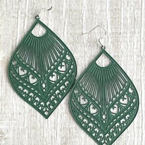 May include: A pair of green, teardrop-shaped earrings with intricate cut-out designs. The earrings feature a fan-like pattern at the top and a series of heart shapes along the bottom edge. The earrings are suspended from silver-colored ear wires.