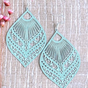May include: A pair of light blue, diamond-shaped drop earrings featuring intricate laser-cut filigree patterns. The design includes radiating lines at the top and heart-shaped cutouts within the lower section, suspended by silver-toned metal hooks.