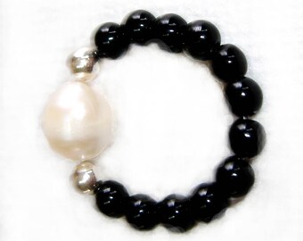 May include: A black beaded ring with a large white pearl. The ring is made of small, round black beads and a single large white pearl.