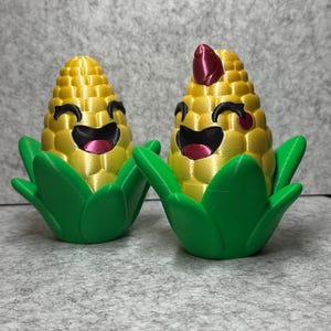 May include: Two 3D printed corn on the cob figurines with smiling faces. The corn is yellow with green husks. One corn has a red top.