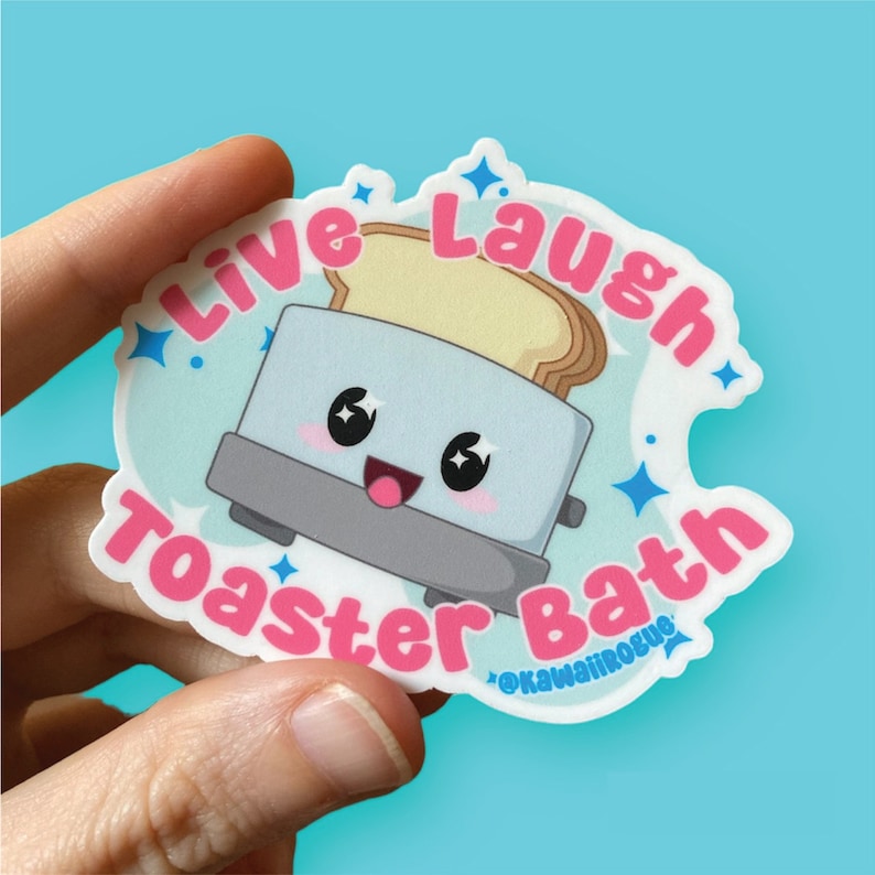 Puede incluir: Una pegatina blanca con una ilustraci&oacute;n de dibujos animados de una tostadora con una rebanada de pan que sale. La tostadora tiene una cara feliz y el texto "Live Laugh Toaster Bath" en letras rosas y azules. El texto "@kawaiirogue" est&aacute; en letras m&aacute;s peque&ntilde;as en la parte inferior.